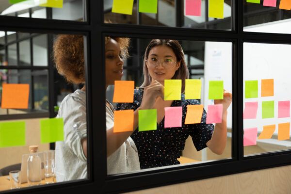 Two women collaborating, standing in front of a window covered with colorful post-it notes, discussing and analyzing ideas together